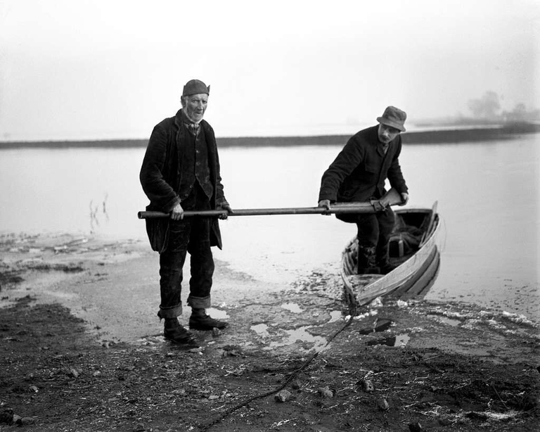 Photo of two men holding a punt gun and stepping out of a boat. 