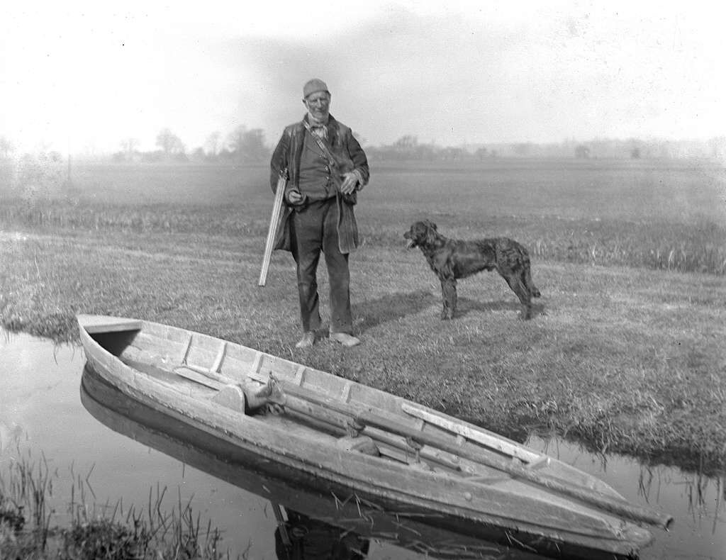 A man holding a rifle while standing next to a boat containing a punt gun.