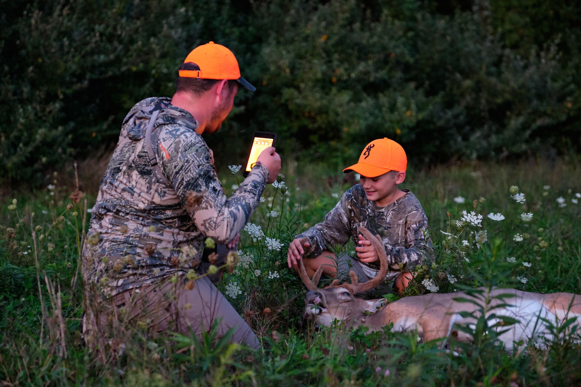 A man and boy in blaze orange hats take a photo with a deer kill, youth hunting safety course concept.