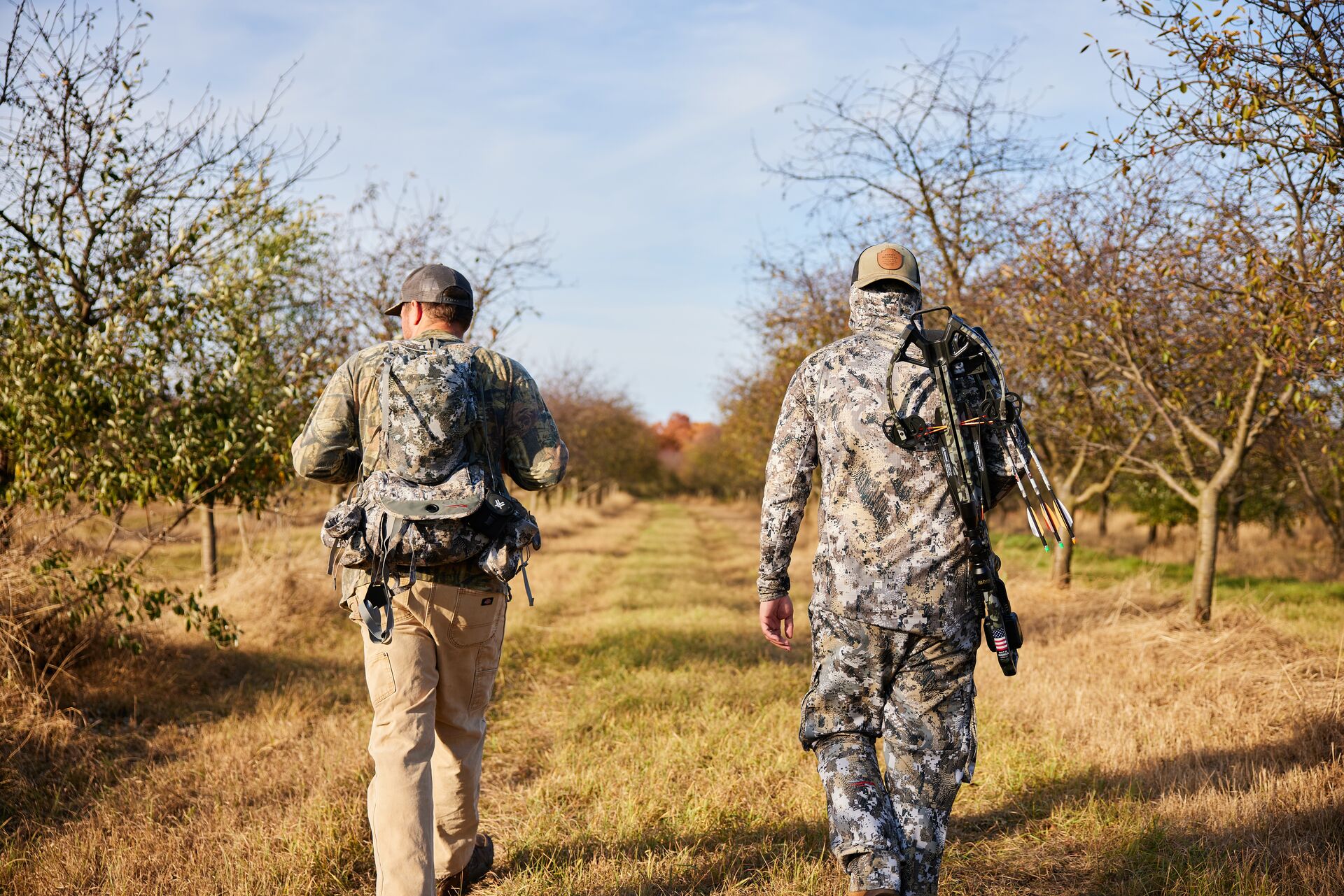 Two hunters walking in a field while wearing camo and carrying hunting gear. 