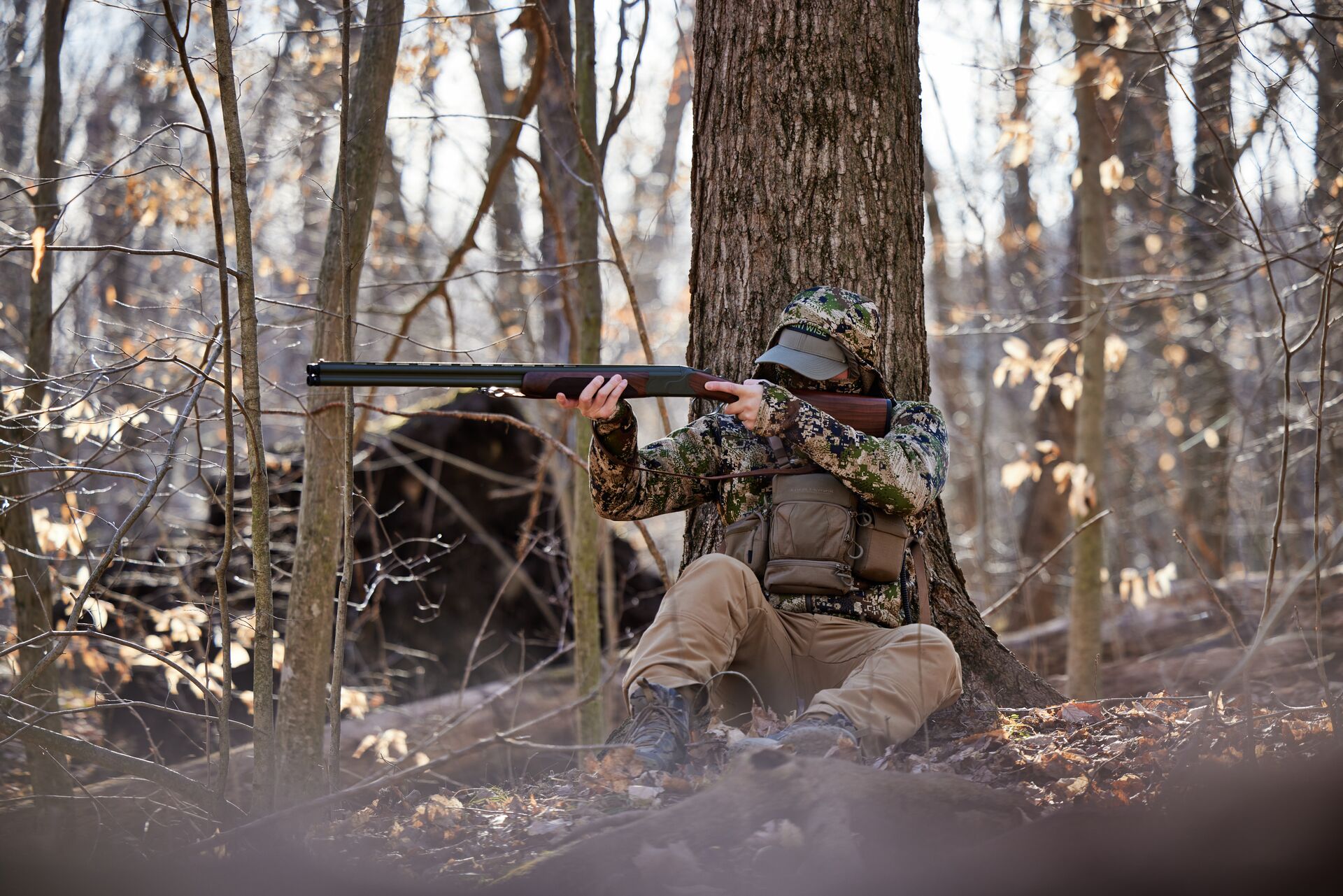 A hunter in camo aims a shotgun while sitting in the woods, hunting turkeys for beginners concept.