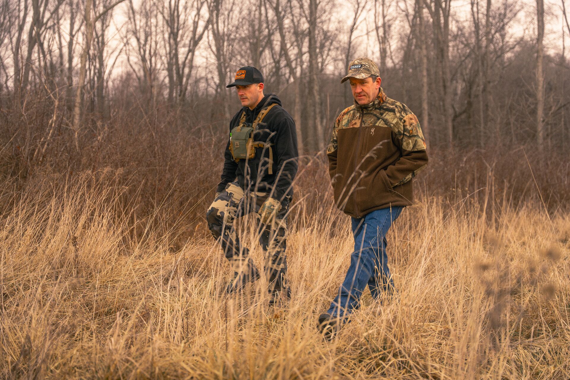 Two hunters walk and talk in a field.