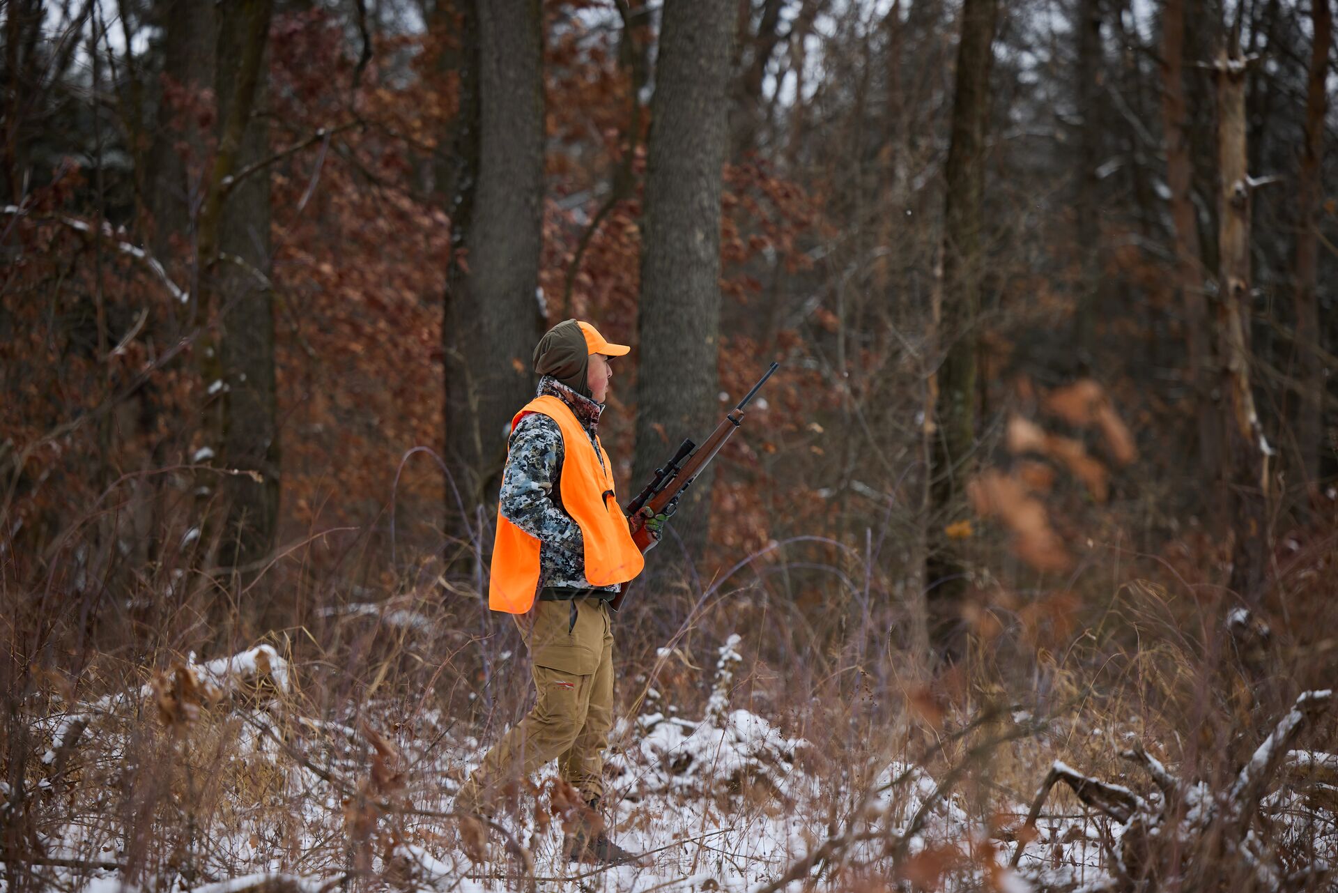Hunter in blaze orange with firearm walks through woods, take a hunter safety course online concept. 